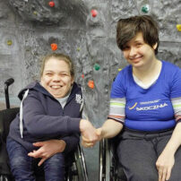 Poniżej znajduje się zdjęcie 2 kobiet z niepełnosprawnością na ściance wspinaczkowej. Siedzą na wózkach.