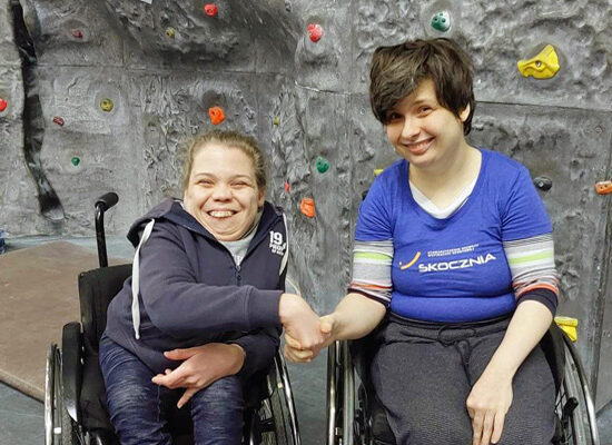 Poniżej znajduje się zdjęcie 2 kobiet z niepełnosprawnością na ściance wspinaczkowej. Siedzą na wózkach.