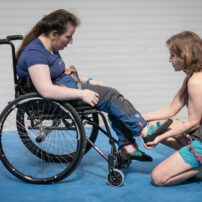 Zdjęcie przedstawia kobietę na wózku z instruktorką wspinania. Obrazuje rozgrzewkę osób z niepełnosprawnością przed wspinaniem