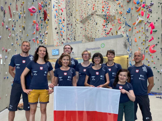 Grupa wspinaczy z niepełnosprawnościami na ściance wspinaczkowej z polską flagą.